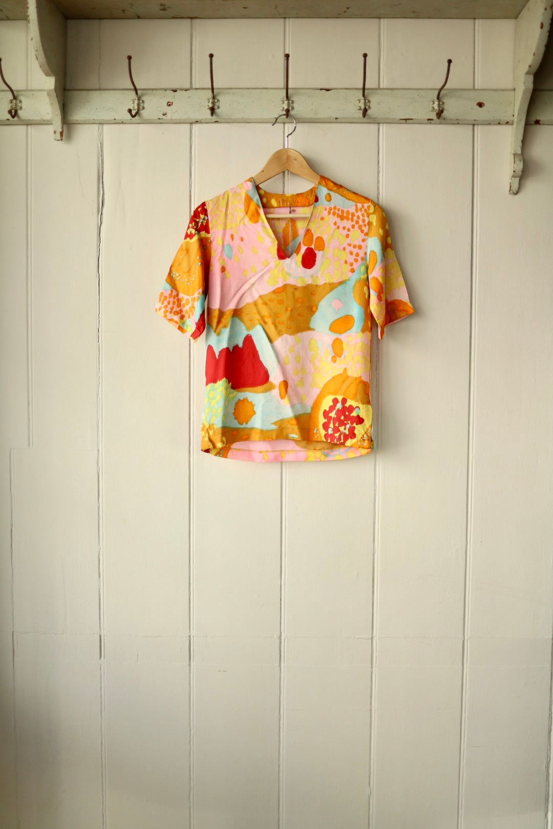 Front view of a short-sleeve top with bold abstract print in warm tones of ochre, red, pink, and aqua, displayed on a wooden hanger against a cream panelled wall.