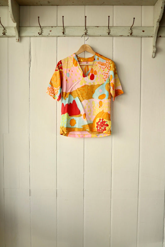 Front view of a short-sleeve top with bold abstract print in warm tones of ochre, red, pink, and aqua, displayed on a wooden hanger against a cream panelled wall.