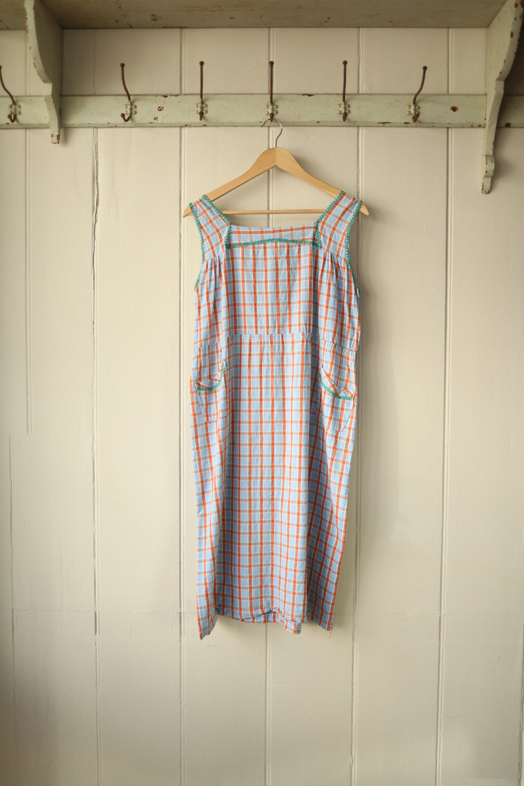 Front view of a vintage home-sewn sleeveless dress in blue, orange and white gingham, with green rik-rak trim on the bust panel, shoulders and patch pockets.
