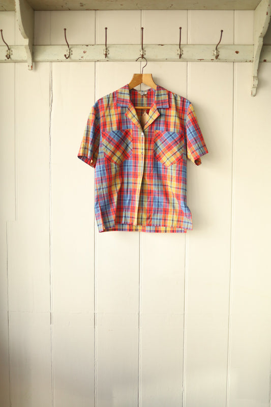 Short-sleeved multicolour plaid shirt with front buttons and two large bias-cut patch pockets, hanging on a wooden hanger against a cream-panelled wall.