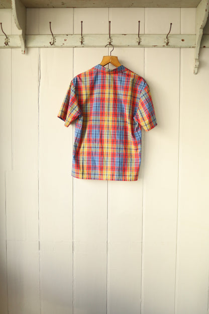 Rear view of the same plaid shirt showing a clean, simple cut in multicoloured check fabric, displayed on a hanger against a cream wall.