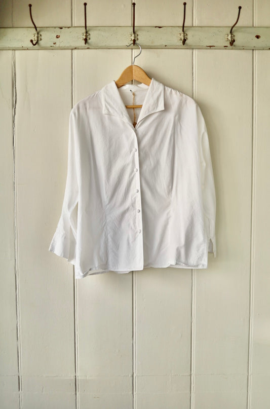 Vintage white blouse with shawl collar and fabric-covered buttons, displayed on wooden hanger against a painted tongue-and-groove wall.