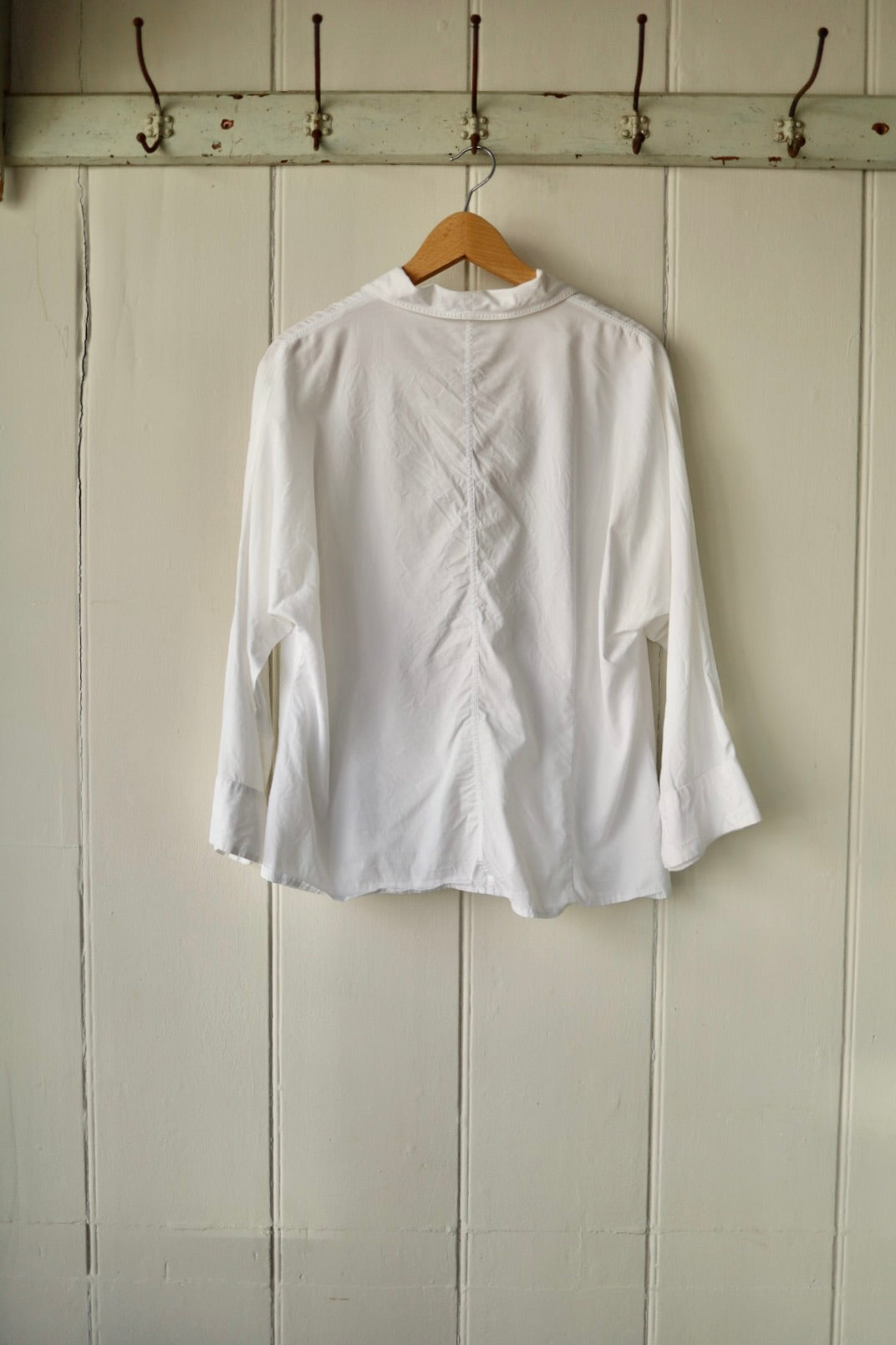 Back of vintage white blouse showing darted seam detailing and flared sleeves, on hanger against panelled cream wall.