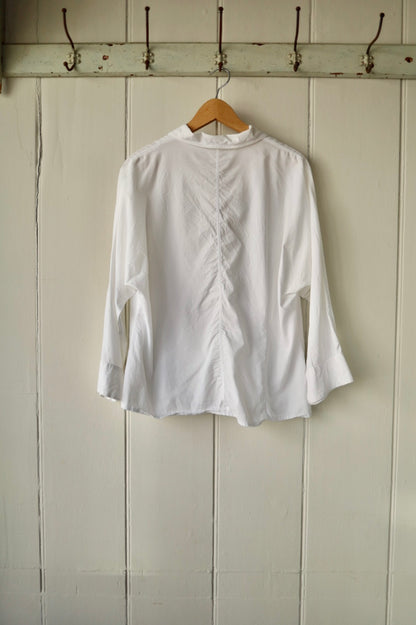 Back of vintage white blouse showing darted seam detailing and flared sleeves, on hanger against panelled cream wall.