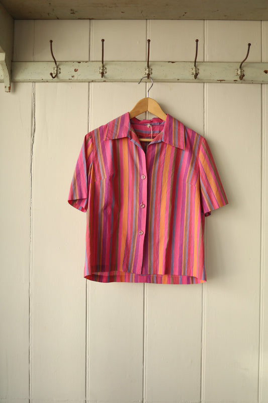 Front view of a short-sleeved button-up shirt in bright candy stripe fabric, featuring vertical stripes in pink, orange, purple, and yellow, hanging on a wooden hanger against a white painted wooden wall with hooks.