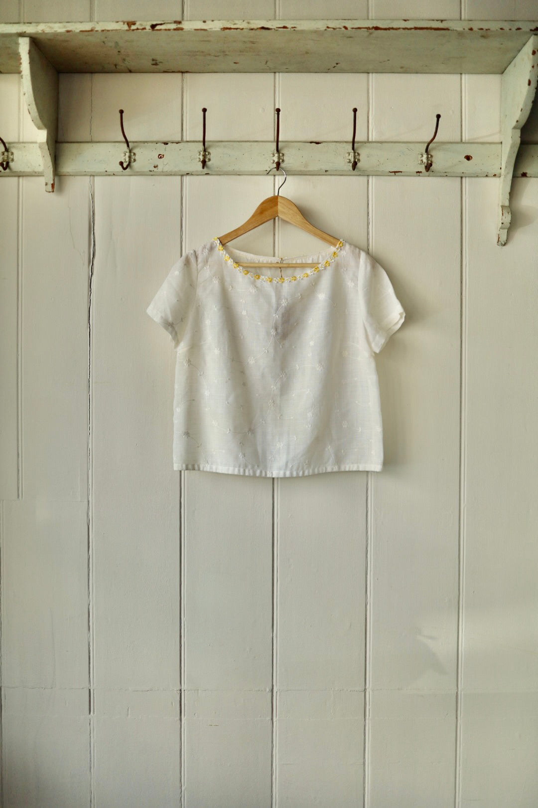 White short-sleeved cotton broderie anglaise top with delicate floral embroidery and a yellow daisy trim along the neckline, displayed on a wooden hanger against a panelled cream wall.