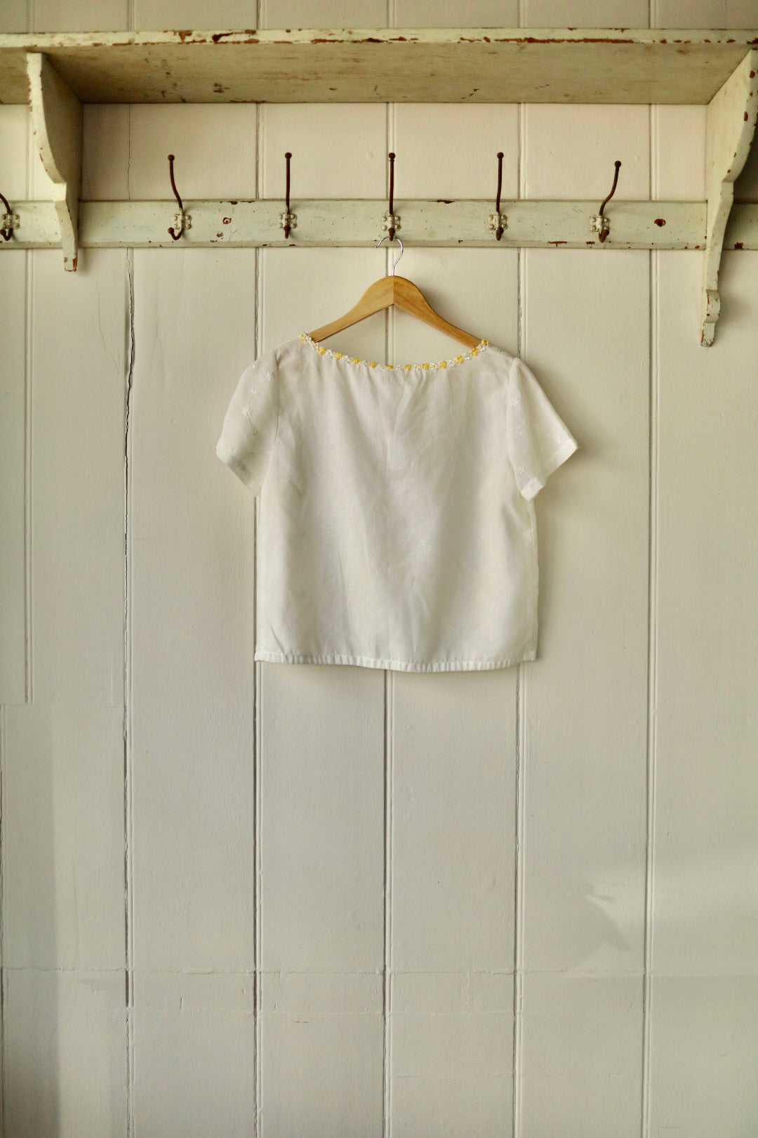 Back view of the same white embroidered cotton top with short sleeves and yellow daisy trim along the neckline, hung neatly on a wooden hanger against a cream panelled wall.