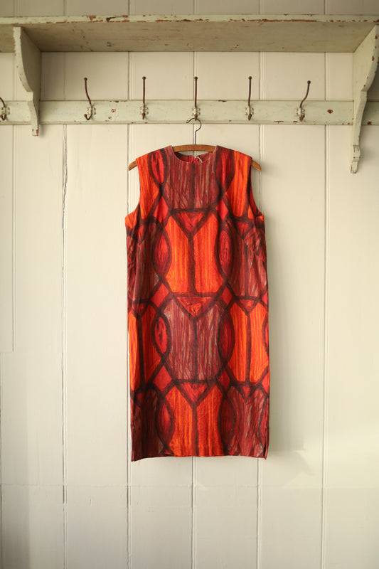 Front view of a sleeveless vintage shift dress in heavy cotton, featuring a bold red and orange abstract flame-like pattern with a high round neckline, photographed against a cream tongue-and-groove wall with a vintage wooden peg rail.