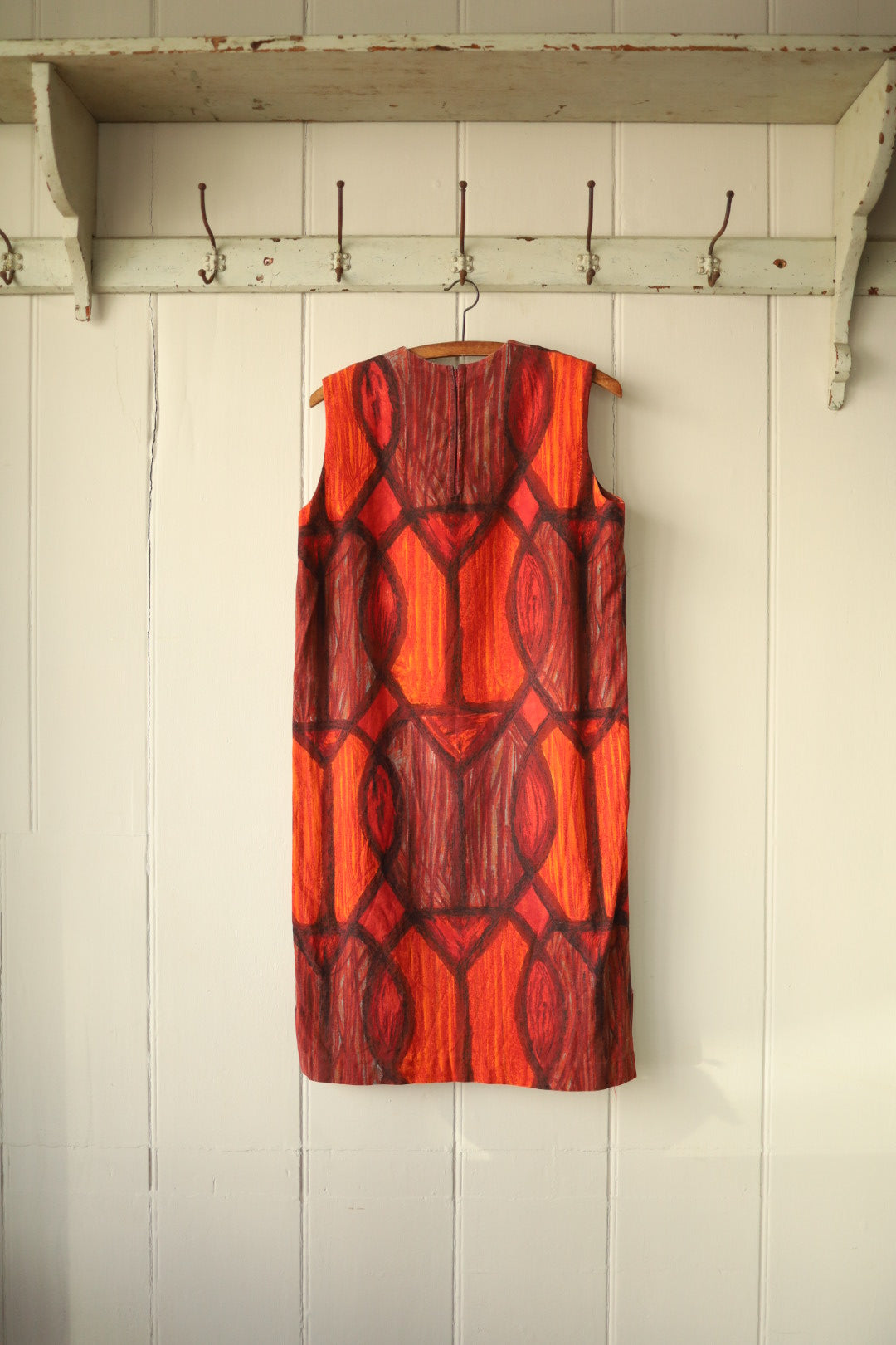 Back view of the same shift dress showing the continuation of the abstract fire print pattern with a concealed zip fastening, hanging on a wooden hanger against a pale panelled wall and rustic peg rail.