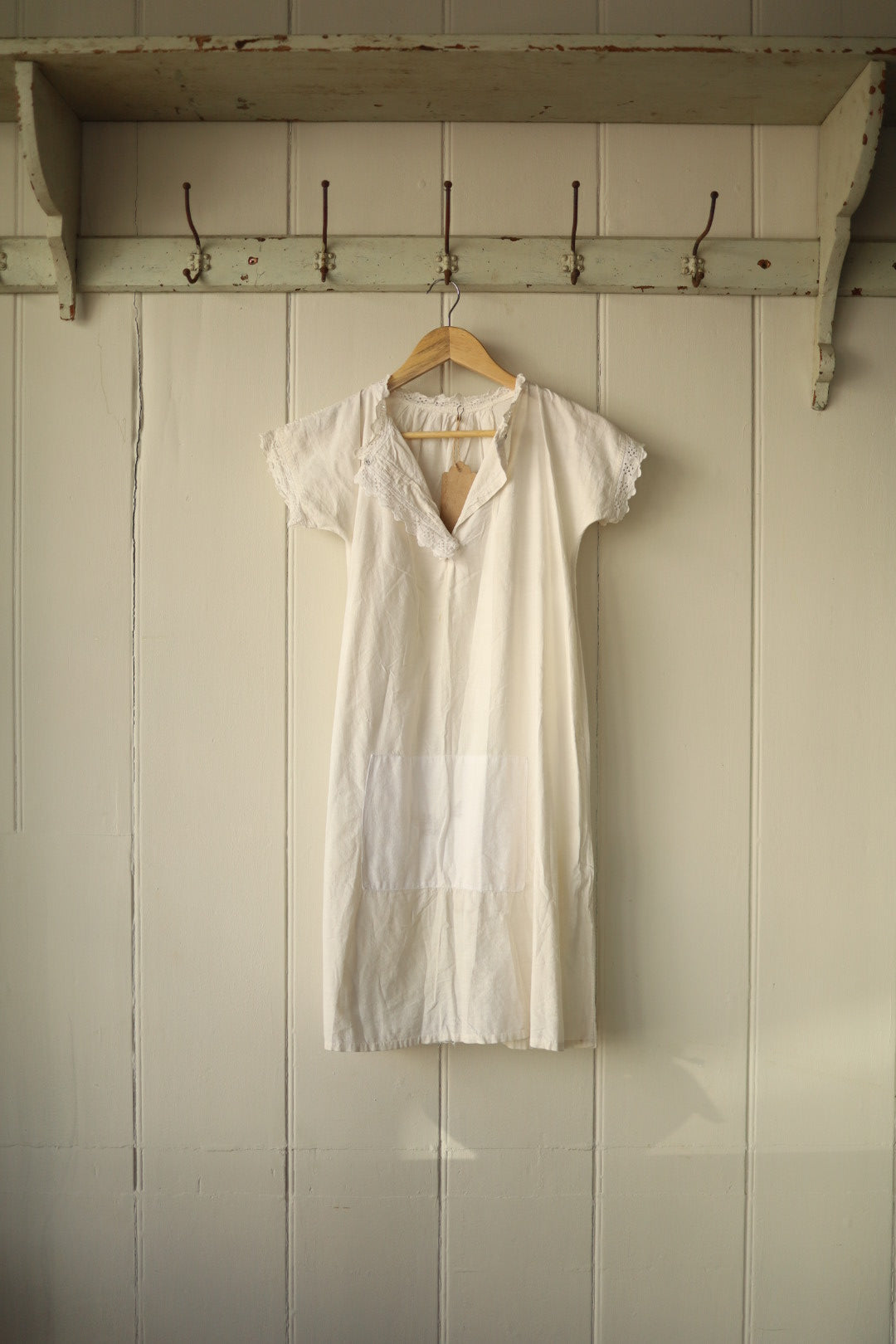White broderie anglaise shirt with lace trim at the collar and sleeve edges, featuring a short sleeve and a large central patch pocket, hung on a wooden hanger against a panelled wall with vintage hooks above.