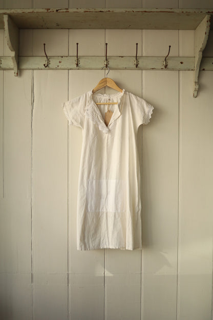 White broderie anglaise shirt with lace trim at the collar and sleeve edges, featuring a short sleeve and a large central patch pocket, hung on a wooden hanger against a panelled wall with vintage hooks above.