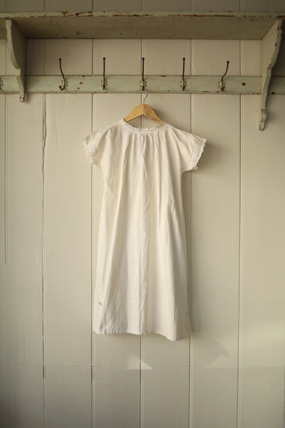 Back of the same white broderie anglaise shirt, showing subtle gathering at the top and lace-trimmed sleeves, displayed on a wooden hanger against the same panelled background.