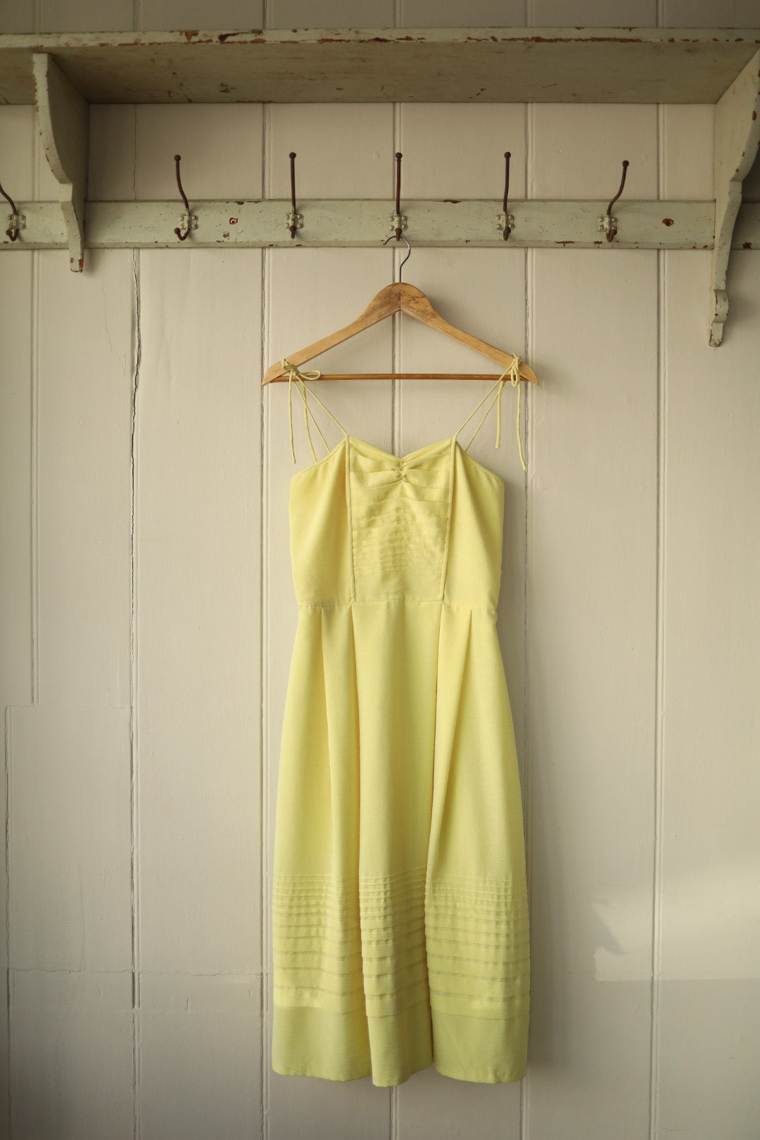 Front view of a vintage spaghetti strap dress in soft sherbet yellow, hanging on a wooden hanger against a white panelled wall. The dress features delicate pintuck detailing on the bodice and a gathered waist that falls into a gently flared skirt.