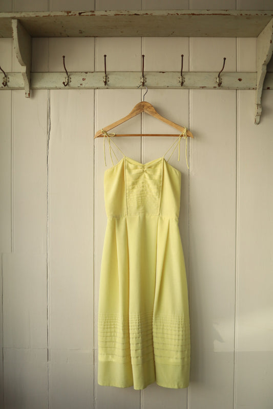 Front view of a vintage spaghetti strap dress in soft sherbet yellow, hanging on a wooden hanger against a white panelled wall. The dress features delicate pintuck detailing on the bodice and a gathered waist that falls into a gently flared skirt.