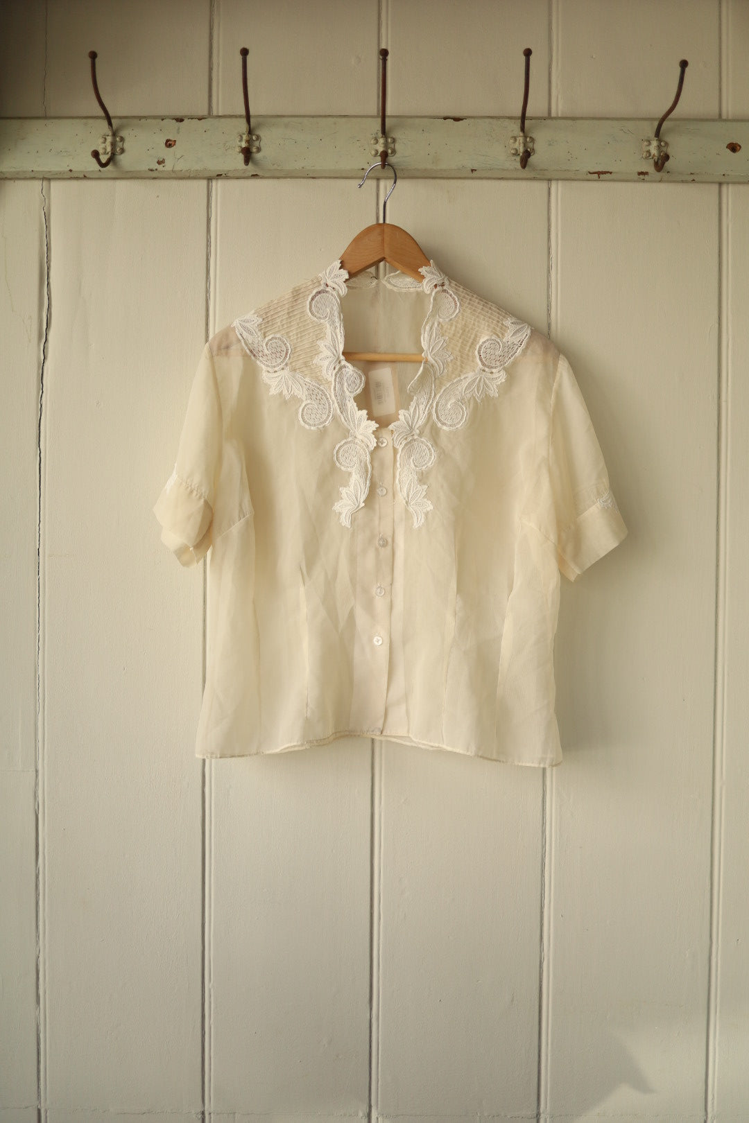 A delicate vintage cutwork blouse in sheer ivory fabric, featuring short sleeves and an ornate floral embroidered collar with scalloped edges, buttoned down the front.
