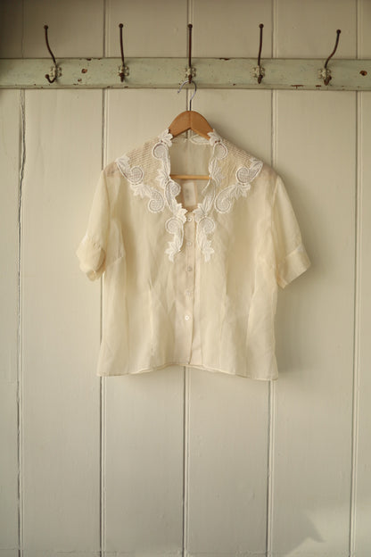 A delicate vintage cutwork blouse in sheer ivory fabric, featuring short sleeves and an ornate floral embroidered collar with scalloped edges, buttoned down the front.