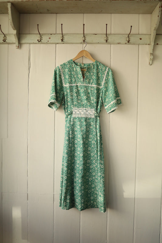 Front view of green ditsy print sun dress with lace trim on bust and waist, hanging on wooden hanger against cream panelled wall