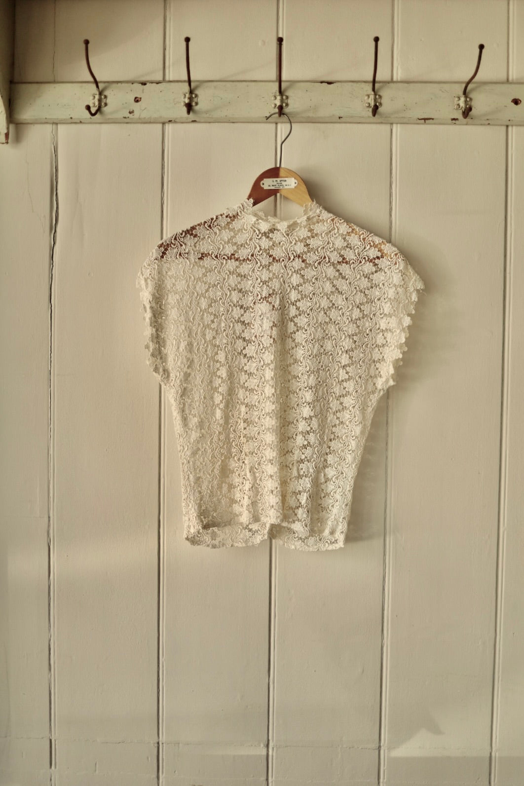 Vintage 1950s Simon Ellis lace blouse in ivory, displayed on a wooden hanger against a panelled cream wall. Intricate floral lace pattern with short sleeves and high neckline.
