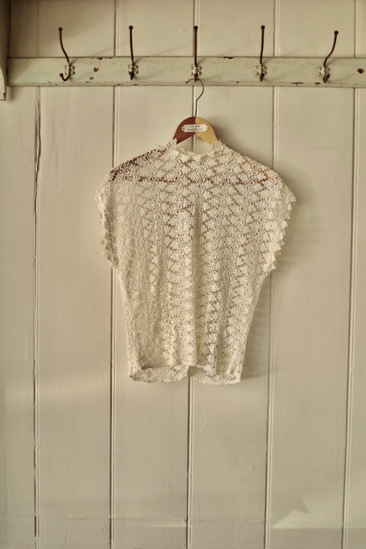 Vintage 1950s Simon Ellis lace blouse in ivory, displayed on a wooden hanger against a panelled cream wall. Intricate floral lace pattern with short sleeves and high neckline.
