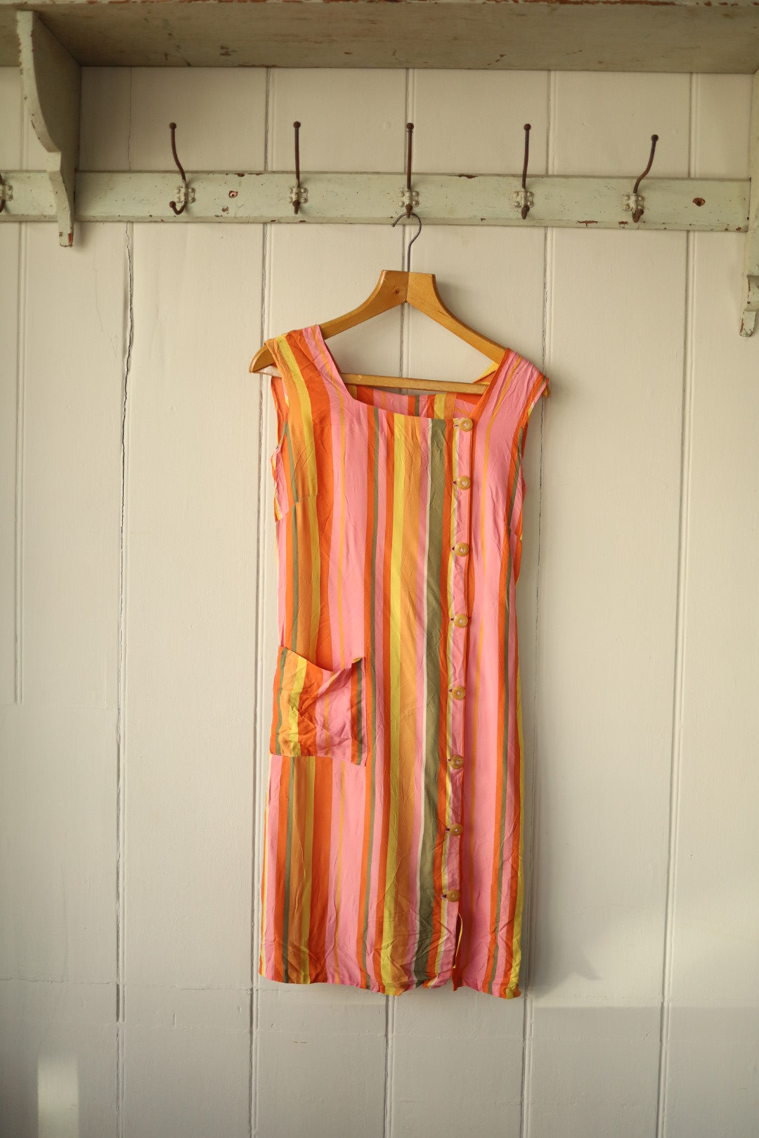 A sleeveless vintage dress in vertical stripes of orange, pink, yellow, and green, with asymmetrical front buttons and a patch pocket on one side, hanging on a wooden hanger against a panelled wall.