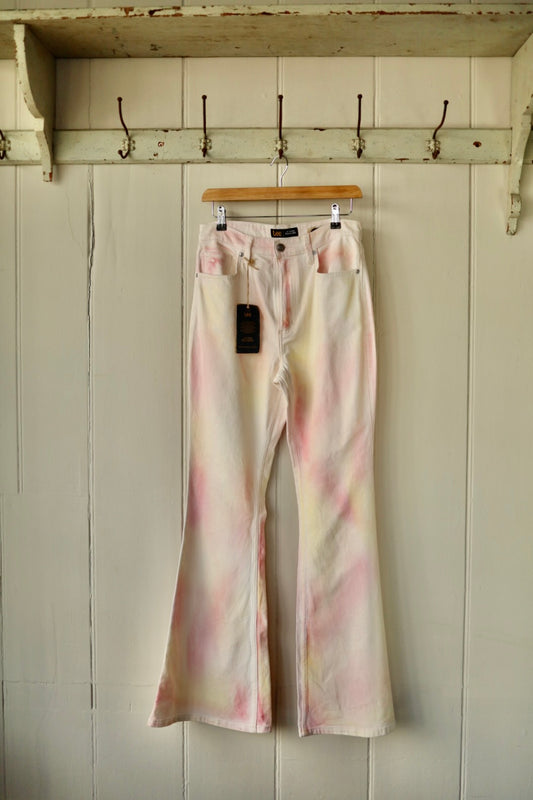 A pair of high-rise Lee flared jeans in a creamy off-white base with soft peach and pink tie-dye effect, shown hanging on a wooden hanger against a cream painted panel wall.