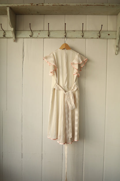 Rear view of the same cream dress showing flutter sleeves, concealed back zip, and long sash tie in matching fabric for a cinched waist.