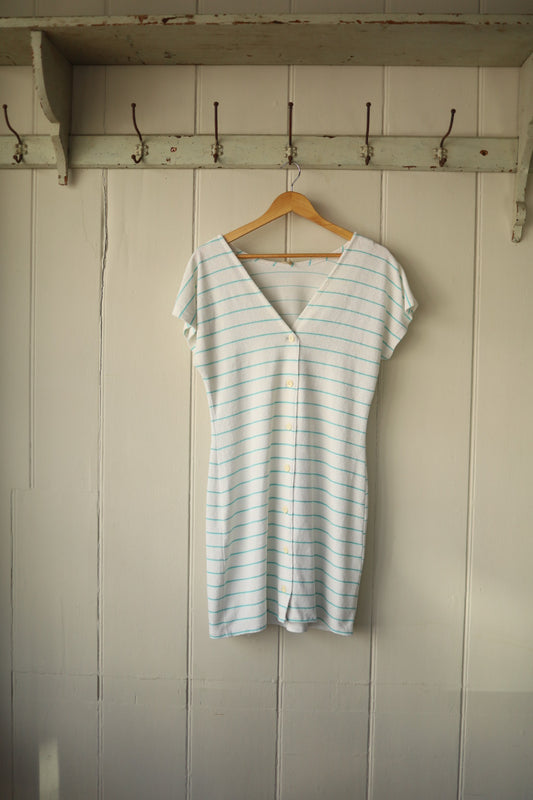 Front view of a short-sleeved towelling beach dress with turquoise horizontal stripes and a V-neckline, displayed on a wooden hanger against a painted wooden wall.