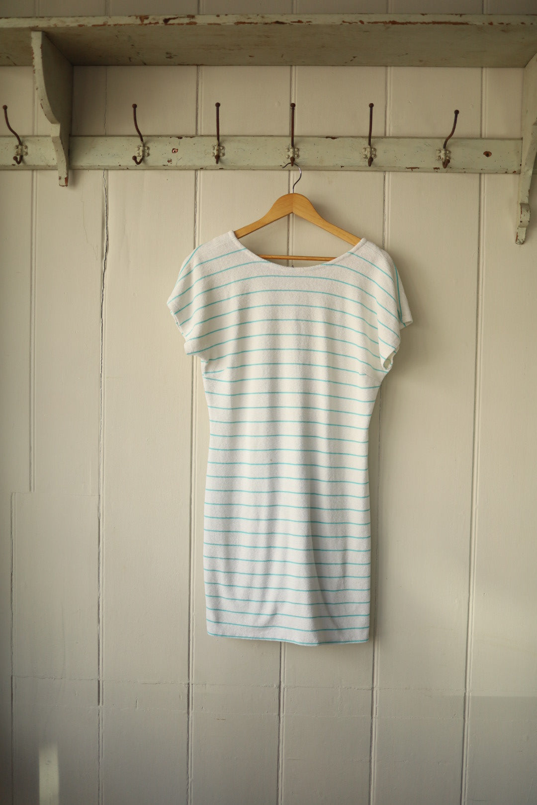 Back view of the same turquoise-striped towelling beach dress, showing a gently scooped back neckline and a straight silhouette, hung on a wooden hanger.