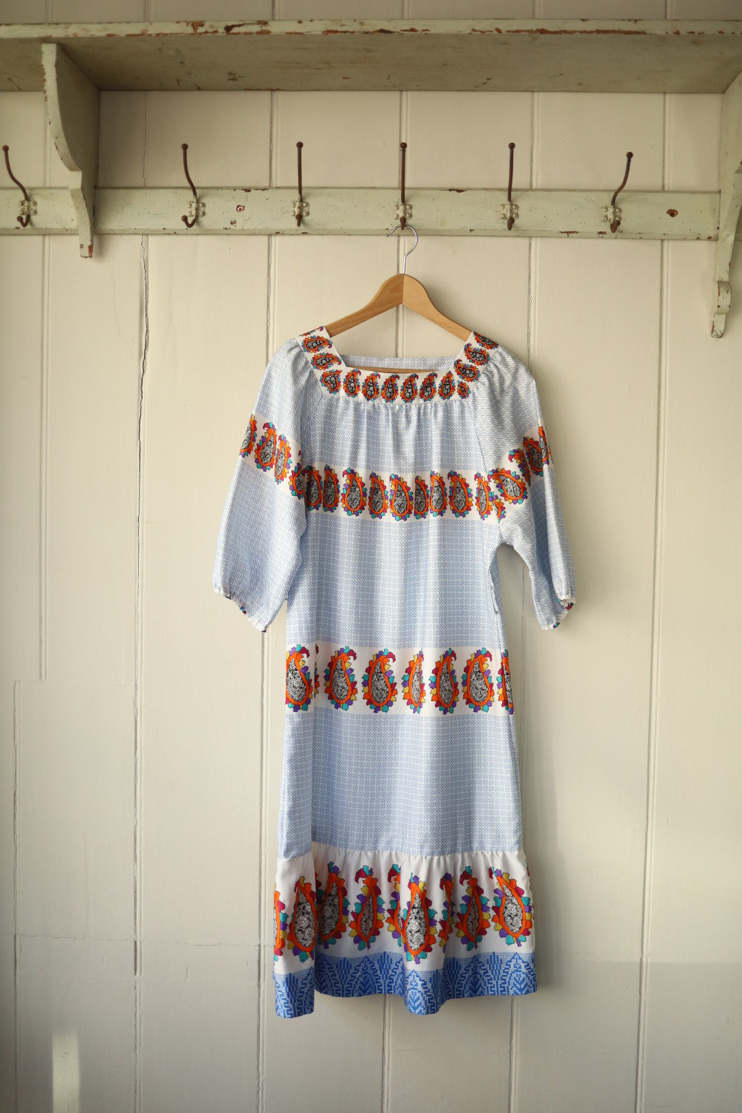 A square-necked vintage dress with puffed sleeves, featuring a blue and white micro-check pattern and vibrant orange paisley print in horizontal bands. The dress hangs on a wooden hanger against a pale panelled wall with hooks above.