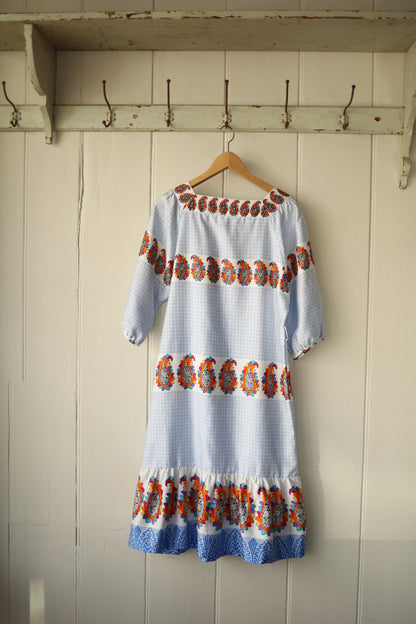 Back view of the same dress, showing its symmetrical paisley banding across the shoulders, sleeves, and hemline, with the gathered square neckline and a gently flared skirt. The dress is displayed on a hanger in natural light against a rustic interior.