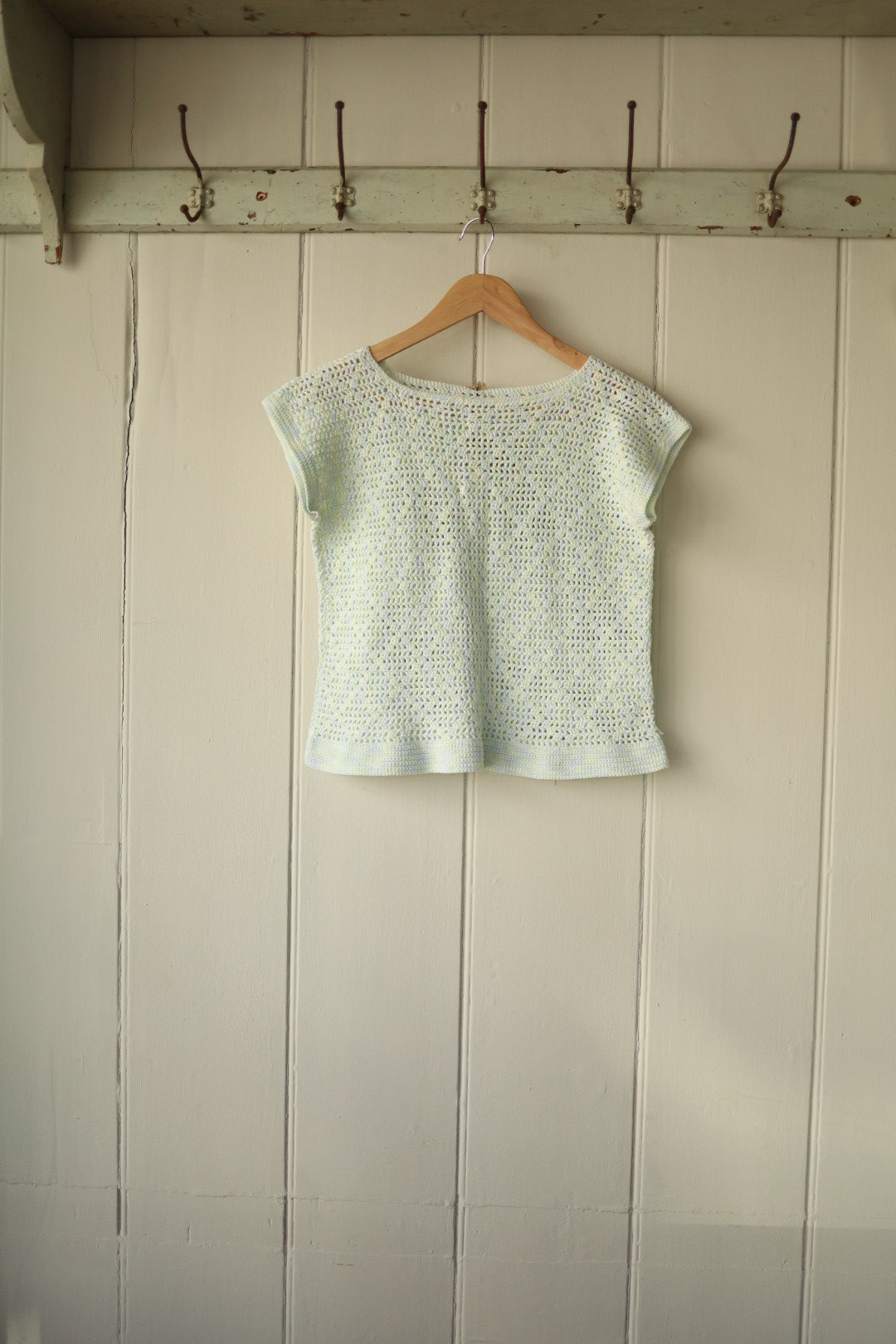Hand-crocheted cropped top in pale blue, featuring a delicate open stitch pattern and cap sleeves, displayed hanging against a panelled wall.