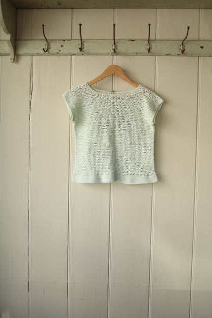 Hand-crocheted cropped top in pale blue, featuring a delicate open stitch pattern and cap sleeves, displayed hanging against a panelled wall.