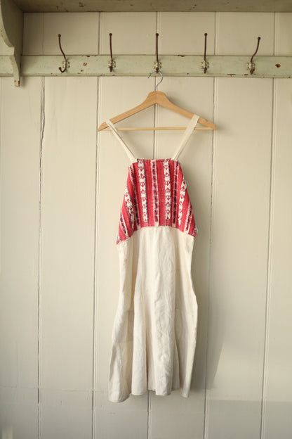  A simple folk dress with a woven red and cream patterned bust and cream skirt, hanging on a wooden hanger against a painted wooden wall.