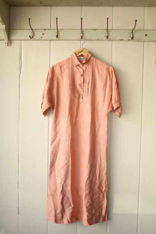 Vintage nude silk dress hanging on a wooden hanger against a white panelled wall, featuring a bib front with pleated detailing and button fastening at the collar.