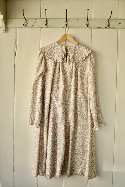 Front view of a vintage-style smock dress with a prairie collar, long sleeves, and a soft floral print in muted pastel tones, hanging on a wooden hanger against a painted panelled wall.