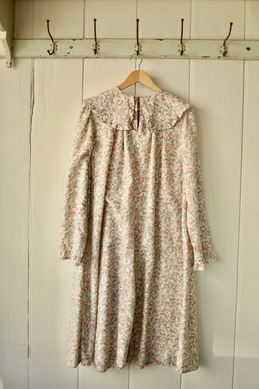 Front view of a vintage-style smock dress with a prairie collar, long sleeves, and a soft floral print in muted pastel tones, hanging on a wooden hanger against a painted panelled wall.