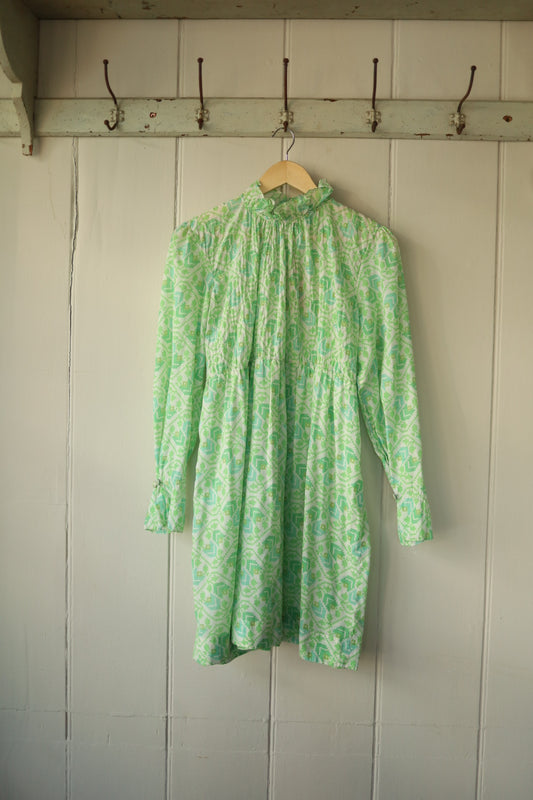 Front view of a vintage smock dress in a bright spring green and white geometric pattern, featuring a high pie crust collar, long sleeves with gathered cuffs, and gentle gathering at the bust.
