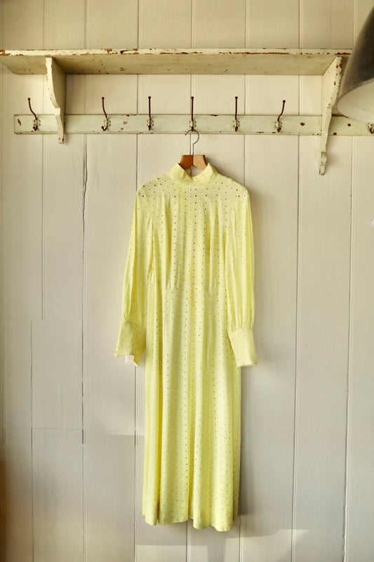 Front view of a vintage yellow broderie anglaise dress with high neck and long sleeves, hanging on a wooden hanger against a panelled wall.