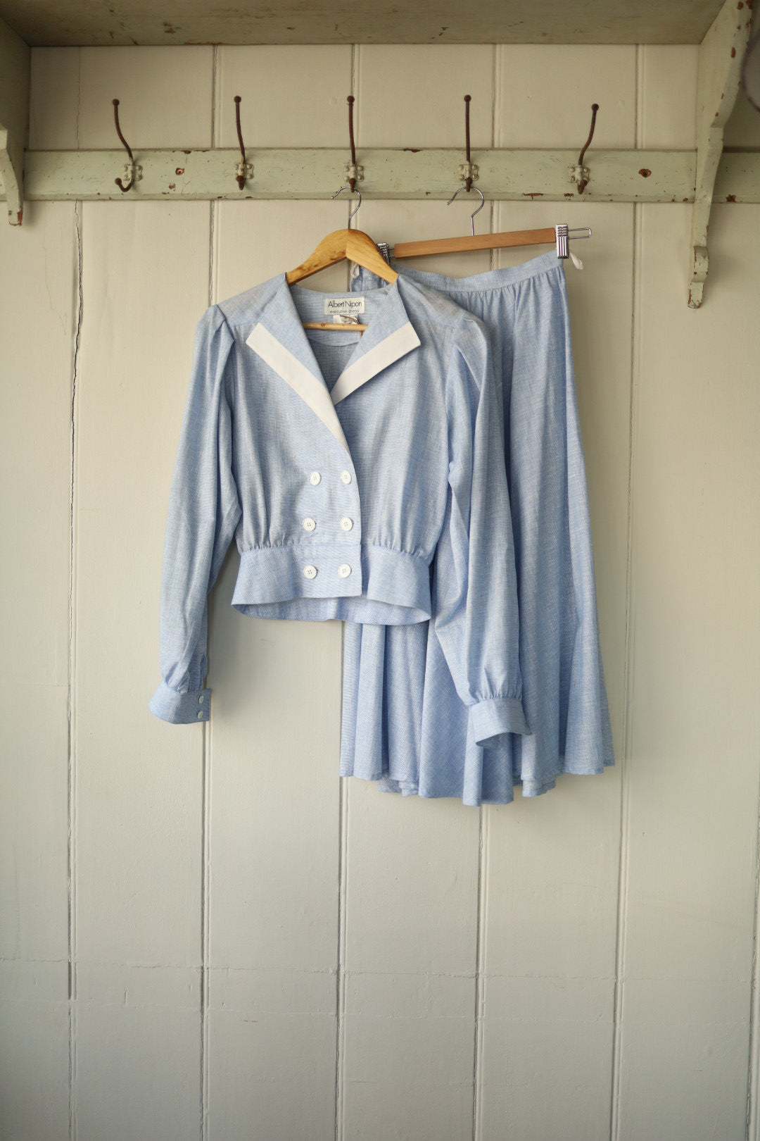 Front view of a light blue Albert Nipon vintage two-piece set with cropped double-breasted jacket and midi skirt, hanging on a wooden hanger against a pale wood-panelled wall.