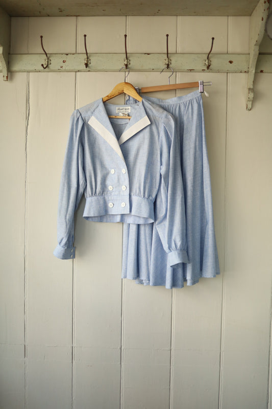 Front view of a light blue Albert Nipon vintage two-piece set with cropped double-breasted jacket and midi skirt, hanging on a wooden hanger against a pale wood-panelled wall.