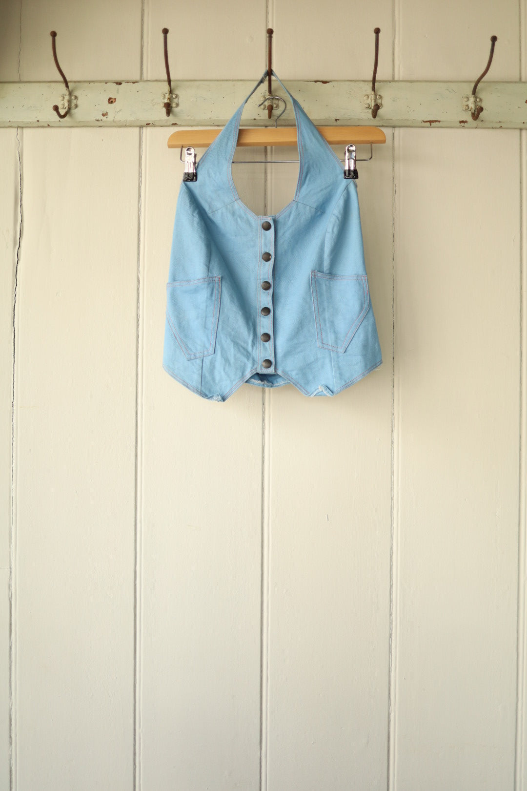 A 70s-style halterneck top in denim-look cotton, featuring a pointed hem and front button fastening, hung on a wooden hanger against a neutral panelled wall.