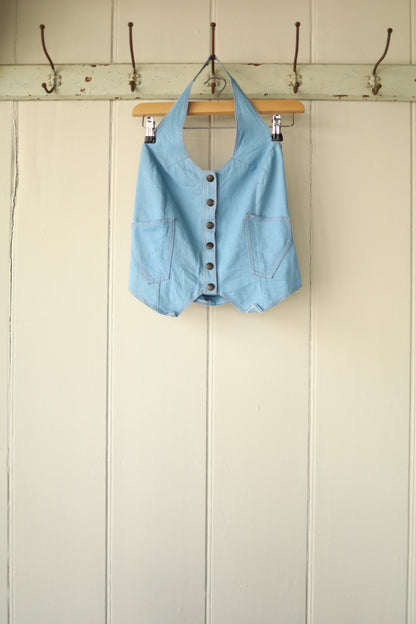 A 70s-style halterneck top in denim-look cotton, featuring a pointed hem and front button fastening, hung on a wooden hanger against a neutral panelled wall.