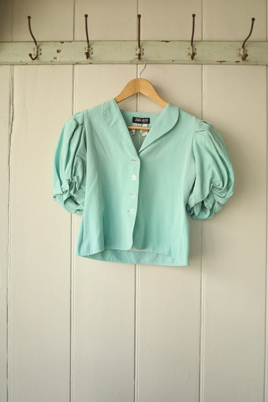 Front view of a Jean Muir mint green short jacket with puffed sleeves and fabric-covered buttons, hanging on a wooden hanger.