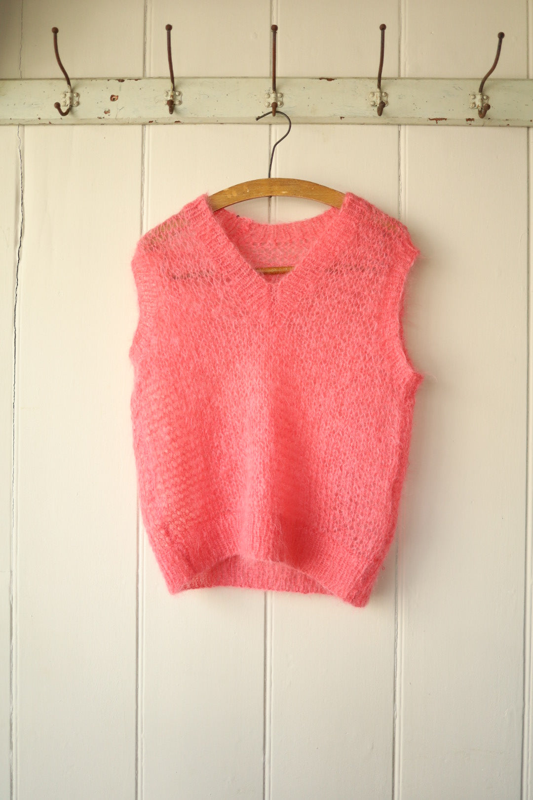 Front view of a hand-knitted bubblegum pink mohair vest hanging on a wooden hanger against a panelled wall.