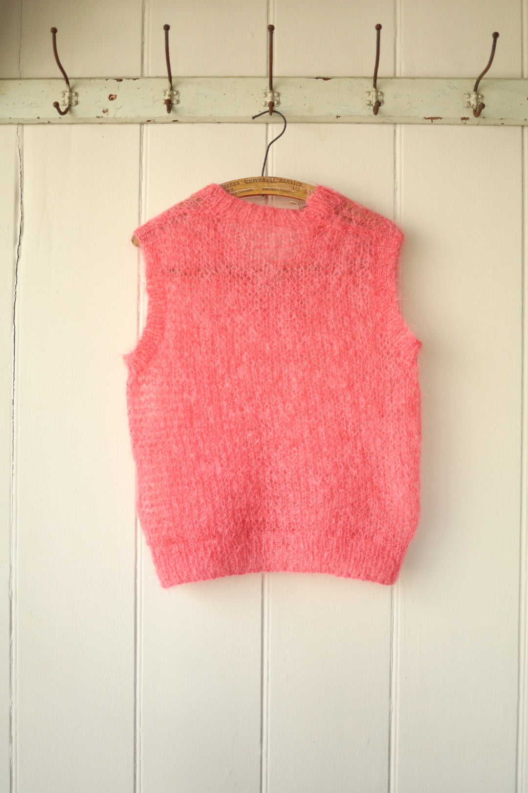 Front view of a hand-knitted bubblegum pink mohair vest hanging on a wooden hanger against a panelled wall.