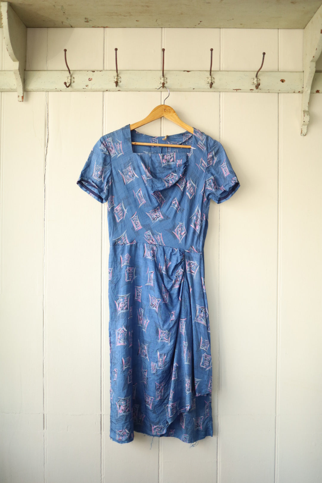 Front view of a vintage blue shantung dress with geometric pink and purple print, short sleeves, and draped skirt, hanging on a wooden hanger against a panelled wall.