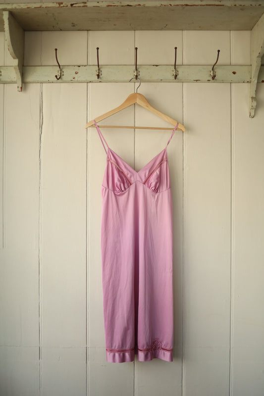 Front view of a raspberry pink vintage satin petticoat dress with stitched bust details, hanging on a wooden hanger against a cream panel wall.