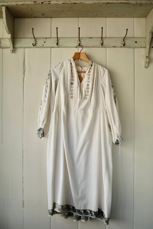 Cream cotton folk dress with embroidered sleeves and hem, gathered neckline, and voluminous silhouette, hanging on a wooden hanger against a pale panelled wall.