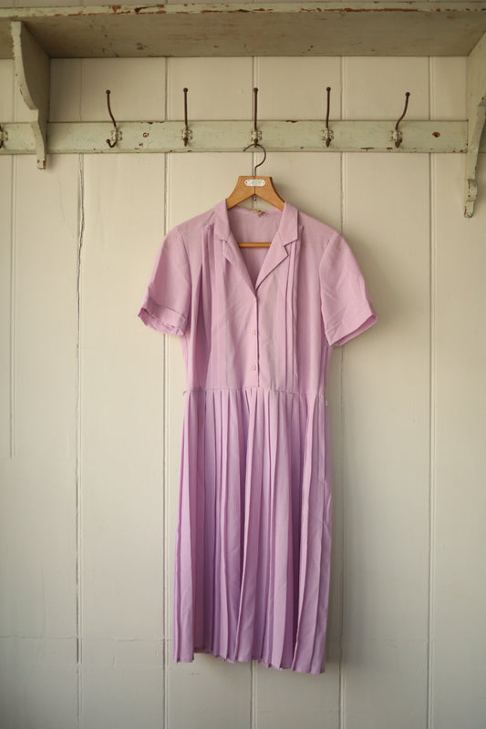 A vintage short-sleeved dress in soft lilac with a buttoned bodice, collar, and pleated skirt, hanging on a wooden hanger against a pale panelled wall.