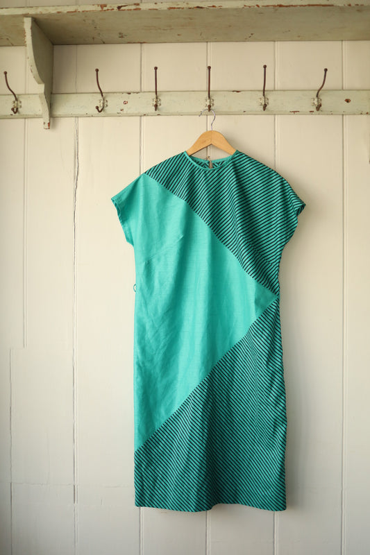 A turquoise and black striped shift dress with asymmetric diagonal panel detailing, displayed on a wooden hanger against a cream-panelled wall.